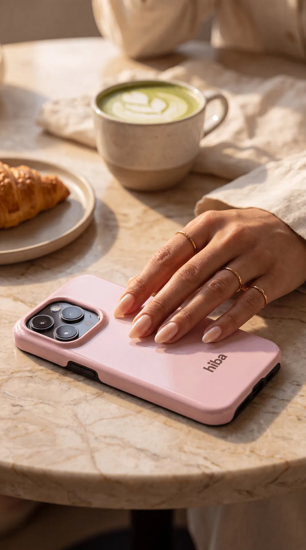 A hand in gold rings resting on a blush HIBA case beside a matcha latte and croissant